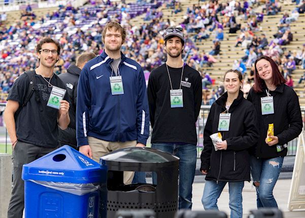 Five students smiling at a football game standing in front of garbage and recycling bins.